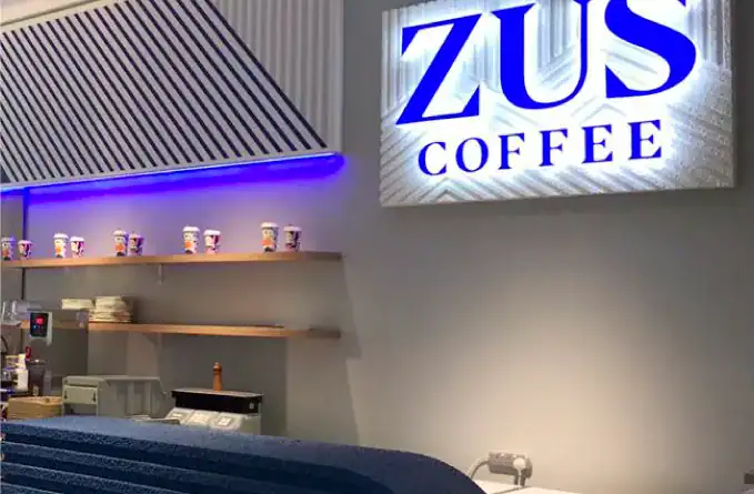 Modern coffee shop interior with ZUS Coffee sign illuminated, stylish decor, display shelves with coffee cups, and customer counter setup in Malaysia for franchise opportunities.